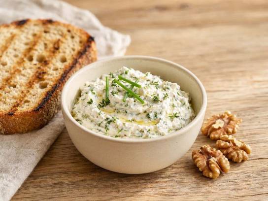 Tartinade de chèvre frais aux herbes dans un bol en céramique avec noix et pain grillé sur une table en bois.
