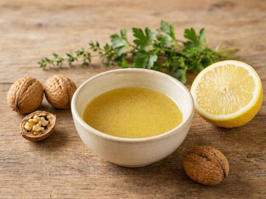 Vinaigrette au citron et à l'huile de noix dans un bol en céramique, entourée de noix et herbes sur table en bois.