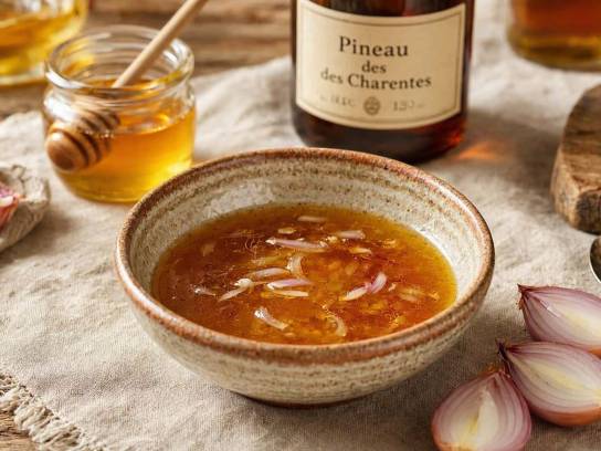 Vinaigrette ambrée au Pineau des Charentes et au miel dans un bol en céramique rustique, entourée d'échalotes fraîches sur une nappe en lin.